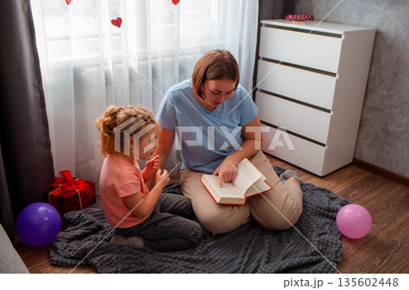 Mid shot of teacher is reading book to a little girl, sitting on the floor. The concept of preschool education for children and Mother's Day	 135602448