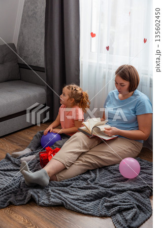 Wide shot of Caucasian little happy girl and her mother sits on carpet in room and reads book. Woman and her daughter spend time. Vertical. Mother's Day Concept Wide shot of Caucasian little happy girl and her mother sits on carpet in room and reads book. Woman and her daughter spend time. Vertical. Mother's Day Concept 135602450