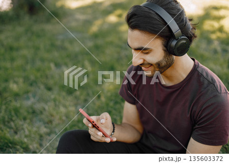 Young boy listening to music while sitting on a grass. Man using a smartphone. Man wearing a bordo shirt and jeans. Young boy listening to music while sitting on a grass. Man using a smartphone. Man wearing a bordo shirt and jeans. 135603372