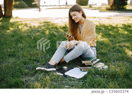 Young girl using a smartphone while sitting on a grass. Woman have a books around her. Girl wearing a beige shirt and jeans. Young girl using a smartphone while sitting on a grass. Woman have a books around her. Girl wearing a beige shirt and jeans. 135603374