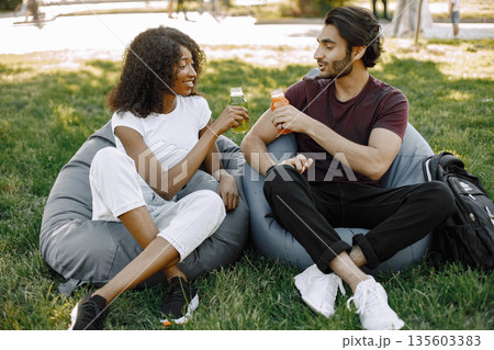 Friends hanging in the green park, relaxing, sitting on the bean bag chairs on grass. Boy and girl holding a bottles of juice. African girl wearing white clothes, boy wearing brown t-shirt and black 135603383