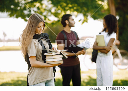 Focus on a caucasian girl who standing sepately. Group of international students standing together in park at university. Blurred african girl and indian boy talking outdoors. 135603399