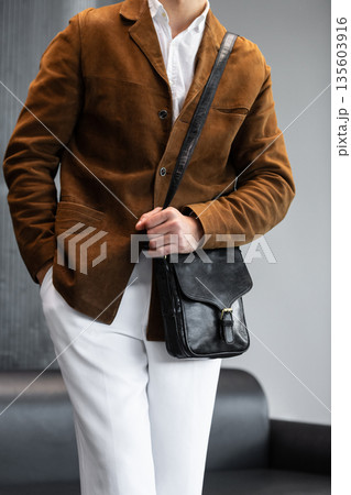 Close-up of a man in a brown corduroy jacket holding a black leather messenger bag with a buckle closure in a minimalist studio. 135603916