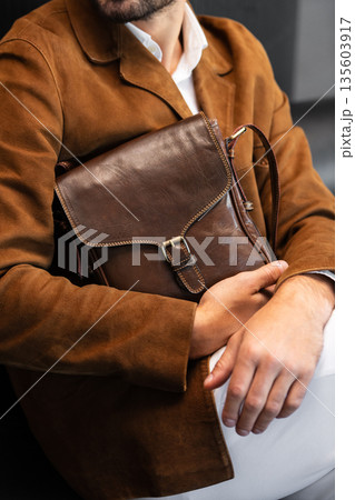 Close-up of a premium leather messenger bag with a metal buckle held by a man in a brown corduroy blazer sitting in a minimalist studio 135603917