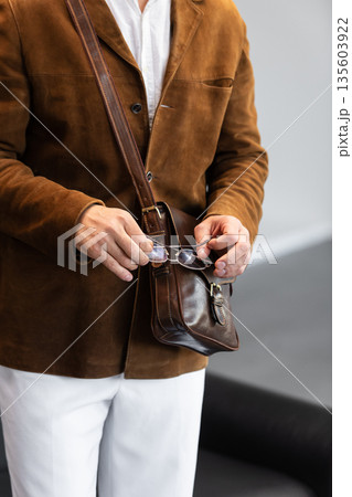 Close-up of a man in a brown corduroy jacket holding a brown leather messenger bag with a buckle closure in a minimalist studio. Close-up of a man in a brown corduroy jacket holding a brown leather messenger bag with a buckle closure in a minimalist studio. 135603922