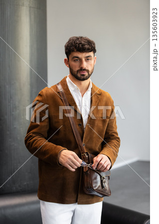 Portrait of a stylish man in a brown corduroy blazer and white shirt holding a cognac leather messenger bag in a minimalist studio. Portrait of a stylish man in a brown corduroy blazer and white shirt holding a cognac leather messenger bag in a minimalist studio. 135603923