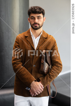 Portrait of a stylish man in a brown corduroy blazer and white shirt holding a cognac leather messenger bag in a minimalist studio. Portrait of a stylish man in a brown corduroy blazer and white shirt holding a cognac leather messenger bag in a minimalist studio. 135603926
