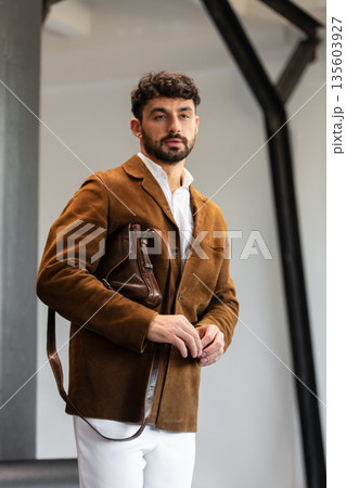 Portrait of a stylish man in a brown corduroy blazer and white shirt holding a cognac leather messenger bag in a minimalist studio. Portrait of a stylish man in a brown corduroy blazer and white shirt holding a cognac leather messenger bag in a minimalist studio. 135603927