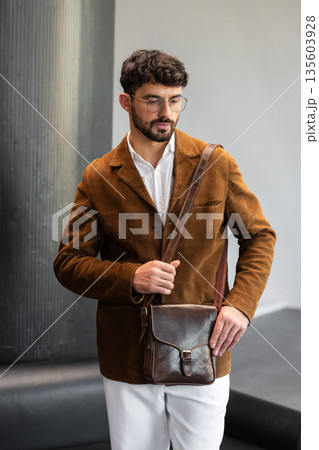 Portrait of a stylish man in a brown corduroy blazer and white shirt holding a cognac leather messenger bag in a minimalist studio. Portrait of a stylish man in a brown corduroy blazer and white shirt holding a cognac leather messenger bag in a minimalist studio. 135603928