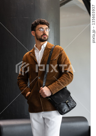 Portrait of a stylish man in a brown corduroy blazer and white shirt holding a black leather messenger bag in a minimalist studio. Portrait of a stylish man in a brown corduroy blazer and white shirt holding a black leather messenger bag in a minimalist studio. 135603937