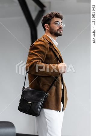 Portrait of a stylish man in a brown corduroy blazer and white shirt holding a black leather messenger bag in a minimalist studio. Portrait of a stylish man in a brown corduroy blazer and white shirt holding a black leather messenger bag in a minimalist studio. 135603943
