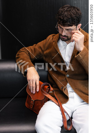 Sophisticated man in a brown corduroy blazer and glasses sitting on a black leather sofa holding a cognac leather messenger bag in a minimalist studio. Sophisticated man in a brown corduroy blazer and glasses sitting on a black leather sofa holding a cognac leather messenger bag in a minimalist studio. 135603950