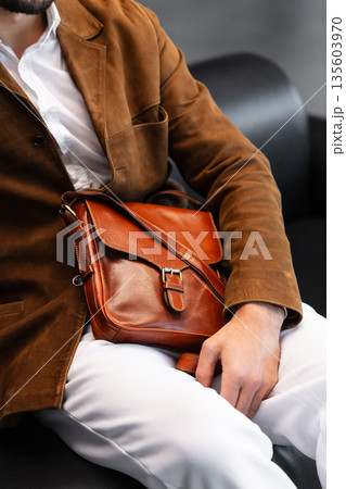 Close-up of a premium cognac leather messenger bag with a metal buckle held by a man in a brown corduroy blazer sitting in a minimalist studio 135603970