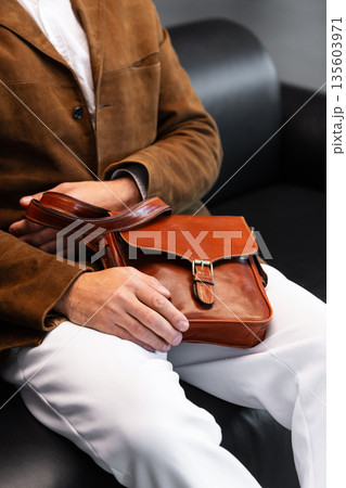 Close-up of a premium cognac leather messenger bag with a metal buckle held by a man in a brown corduroy blazer sitting in a minimalist studio 135603971