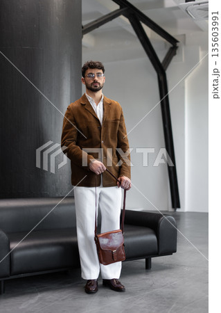 Stylish man in a brown corduroy blazer and white trousers holding a cognac leather messenger bag in a modern minimalist studio interior. Stylish man in a brown corduroy blazer and white trousers holding a cognac leather messenger bag in a modern minimalist studio interior. 135603991
