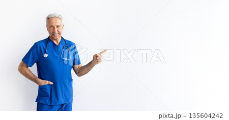 Senior male doctor pointing aside while standing against white studio background Senior male doctor pointing aside while standing against white studio background 135604242