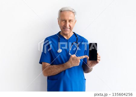 Senior male doctor in blue medical uniform holding smartphone with blank screen 135604256