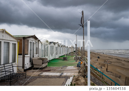 Stormy seaside promenade with closed beach cabins and dramatic dark clouds over empty sandy coast 135604538