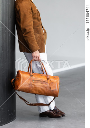Close-up of a man holding a premium brown leather duffel bag wearing a suede blazer and white trousers 135604744
