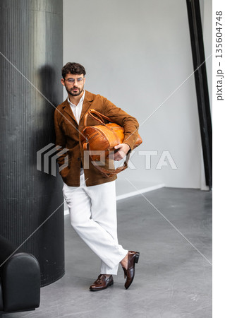 Handsome stylish man in brown suede blazer and white trousers holding premium leather duffel bag in modern interior Handsome stylish man in brown suede blazer and white trousers holding premium leather duffel bag in modern interior 135604748