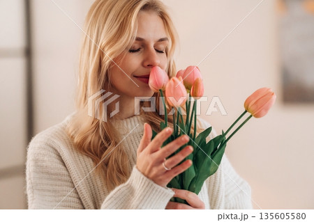 Admiring the gift, standing. Beautiful young woman is at home with flowers Admiring the gift, standing. Beautiful young woman is at home with flowers 135605580