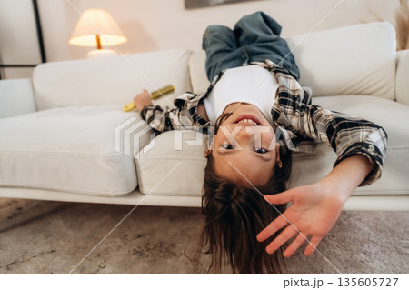 Upside down, lying down on the sofa. Cute little girl is in the domestic room alone Upside down, lying down on the sofa. Cute little girl is in the domestic room alone 135605727