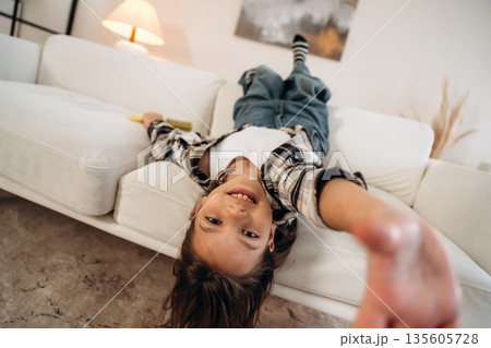 Upside down, lying down on the sofa. Cute little girl is in the domestic room alone Upside down, lying down on the sofa. Cute little girl is in the domestic room alone 135605728