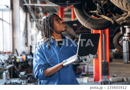 Laptop is in hands. Male mechanic is with car in garage 135605912