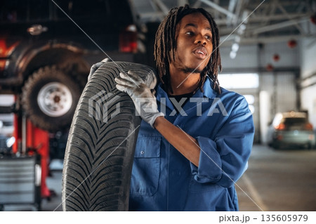 Tire is in hands. Male mechanic is with car in garage Tire is in hands. Male mechanic is with car in garage 135605979