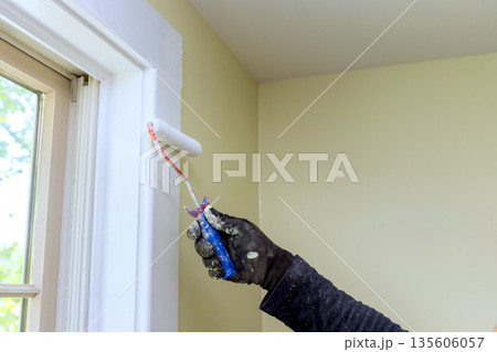 Worker uses small roller to paint wall trim near window in room filled with natural light. 135606057