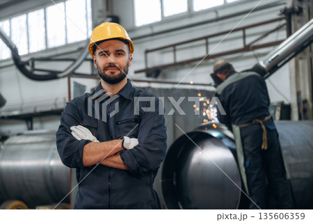 Standing with arms crossed, welding in progress behind. Man is working in the factory 135606359