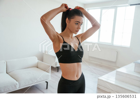 Getting ready for exercises, making ponytail. Young woman with slim body type is indoors Getting ready for exercises, making ponytail. Young woman with slim body type is indoors 135606531