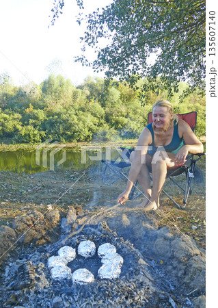Woman bakes potatoes in foil on coals. Woman relaxes in nature. Cooking potatoes Woman bakes potatoes in foil on coals. Woman relaxes in nature. Cooking potatoes 135607140