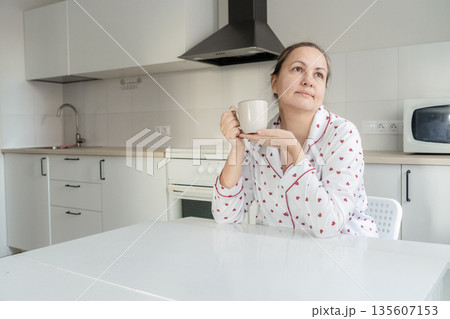 Woman wearing heart print pajamas holding a coffee mug, thinking while sitting at a white table in a bright modern kitchen 135607153