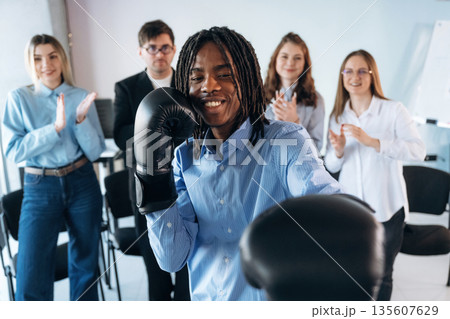 Black man is with boxing gloves. Group of young business people are in conference room Black man is with boxing gloves. Group of young business people are in conference room 135607629