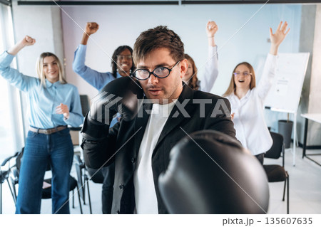Focused guy in glasses is with boxing gloves. Group of young business people are in conference room Focused guy in glasses is with boxing gloves. Group of young business people are in conference room 135607635
