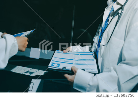 Two doctors and a female nurse meet at a table in the hospital, collaborating on medical tasks using laptops and computers 135608750