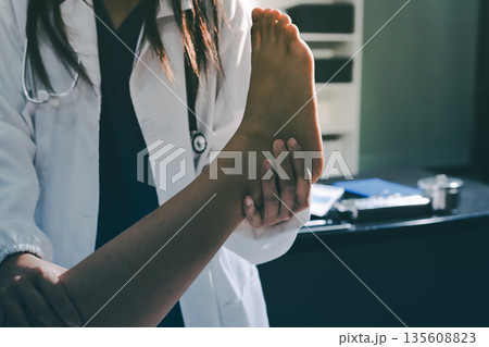 Doctor examining and supporting injured ankle of female patient in hospital room, providing medical assistance and treatment for sprain or fracture 135608823