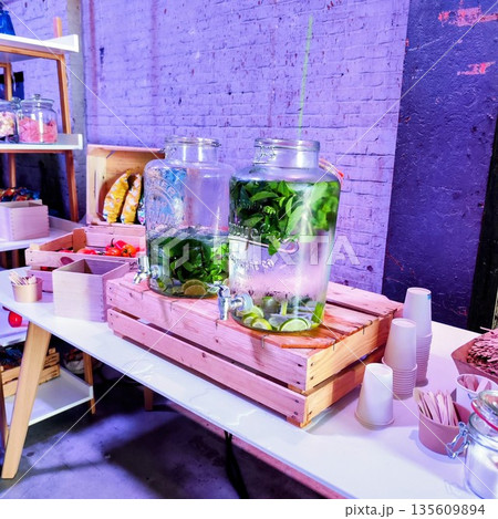 Two large glass jars filled with refreshing beverages are displayed on a table. Each jar contains water infused with lime slices and fresh mint leaves, resting on wooden stands with spigots Two large glass jars filled with refreshing beverages are displayed on a table. Each jar contains water infused with lime slices and fresh mint leaves, resting on wooden stands with spigots 135609894