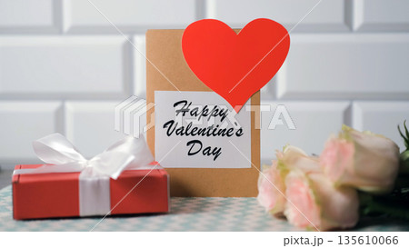 Gift box and Happy Valentines Day card next to a bouquet of roses on the table 135610066