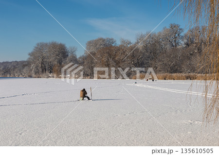 A fisherman sits on a snow-covered frozen river on a clear day A fisherman sits on a snow-covered frozen river on a clear day 135610505