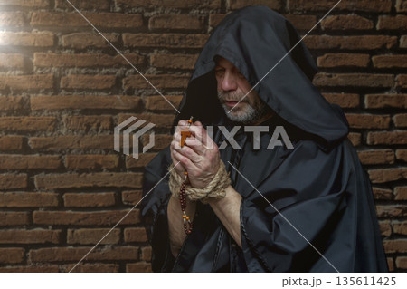 Monk in dark clothing holds rosary with bound hands against old brick wall. Monk in dark clothing holds rosary with bound hands against old brick wall. 135611425