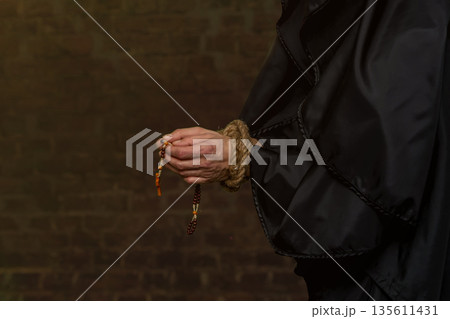 Close-up of a monk's hands tied with rope, holding rosary beads against a blurred dark background. 135611431