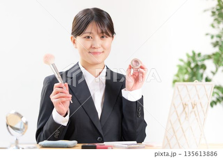 メイク道具を手に持ち、笑顔で紹介するリクルートスーツ姿の女子大学生 メイク道具を手に持ち、笑顔で紹介するリクルートスーツ姿の女子大学生 135613886