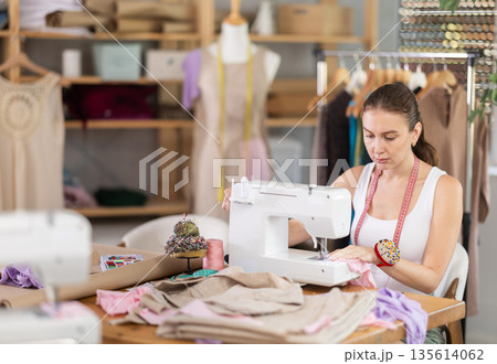 Positive young female dressmaker working with sewing-machine Positive young female dressmaker working with sewing-machine 135614062