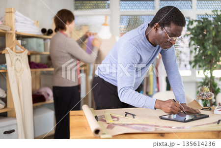 Male tailor draws sketch of future clothing on an electronic tablet. Working with modern gadgets in sewing workshop Male tailor draws sketch of future clothing on an electronic tablet. Working with modern gadgets in sewing workshop 135614236