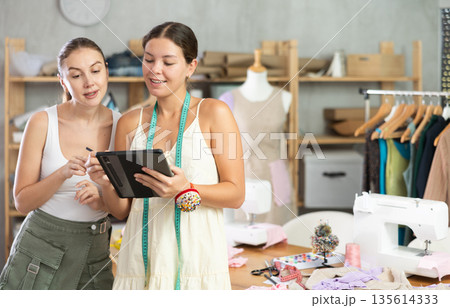 Two female dressmakers drawing a model of clothes on a tablet Two female dressmakers drawing a model of clothes on a tablet 135614333