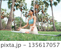 Asian woman meditating in a peaceful park setting, sitting cross-legged on green grass 135617057