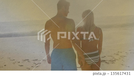 Walking couple wearing swim trunks and bikini on sandy ocean shore toward glowing sunset 135617431