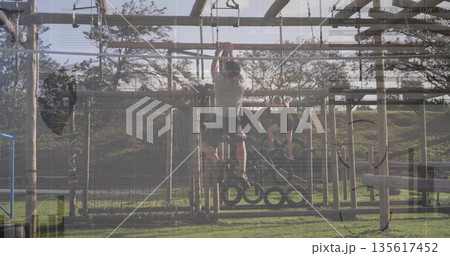 Climbing athletic male swinging on hanging rings in grassy park obstacle course, training partners 135617452